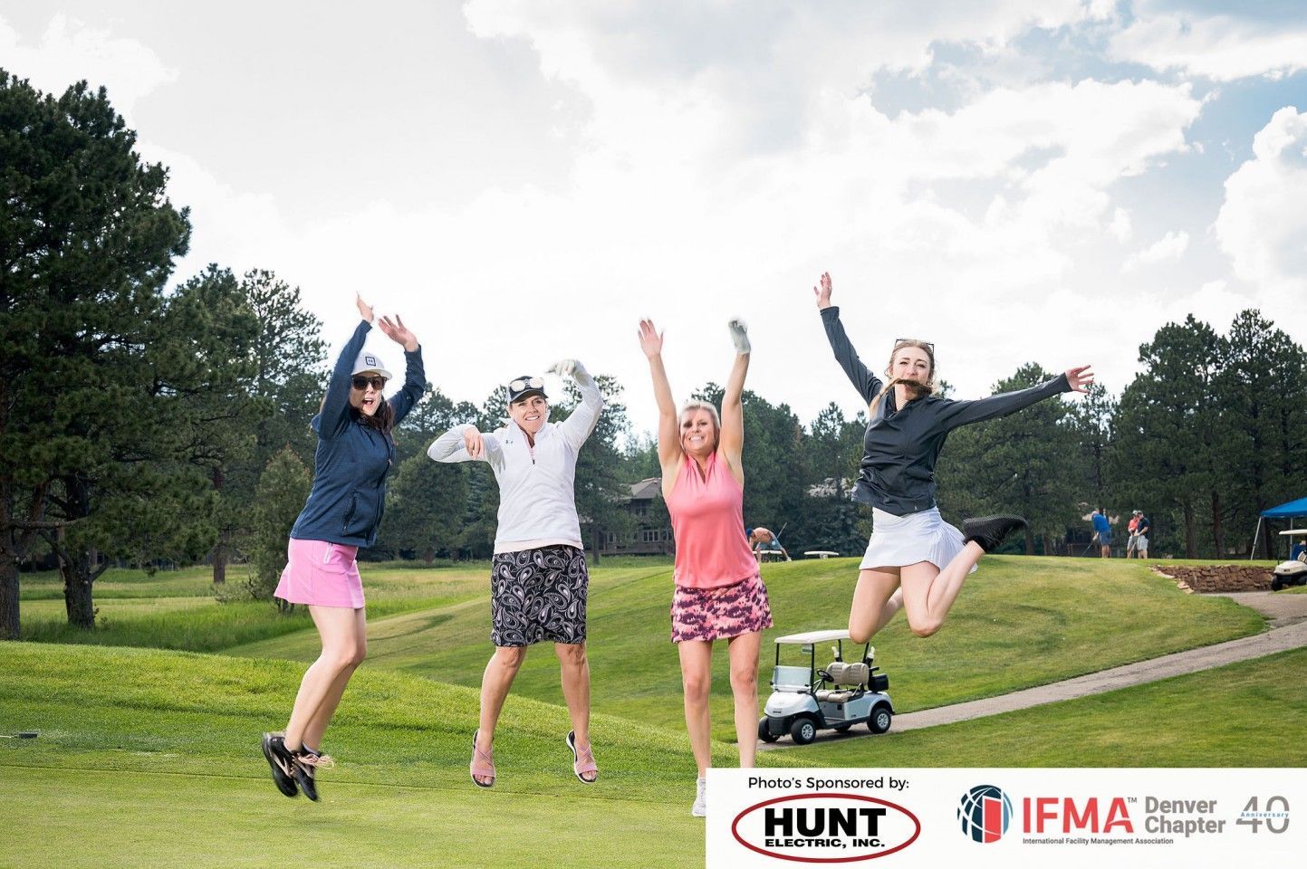 Four women in golf attire jump for joy on a golf course.