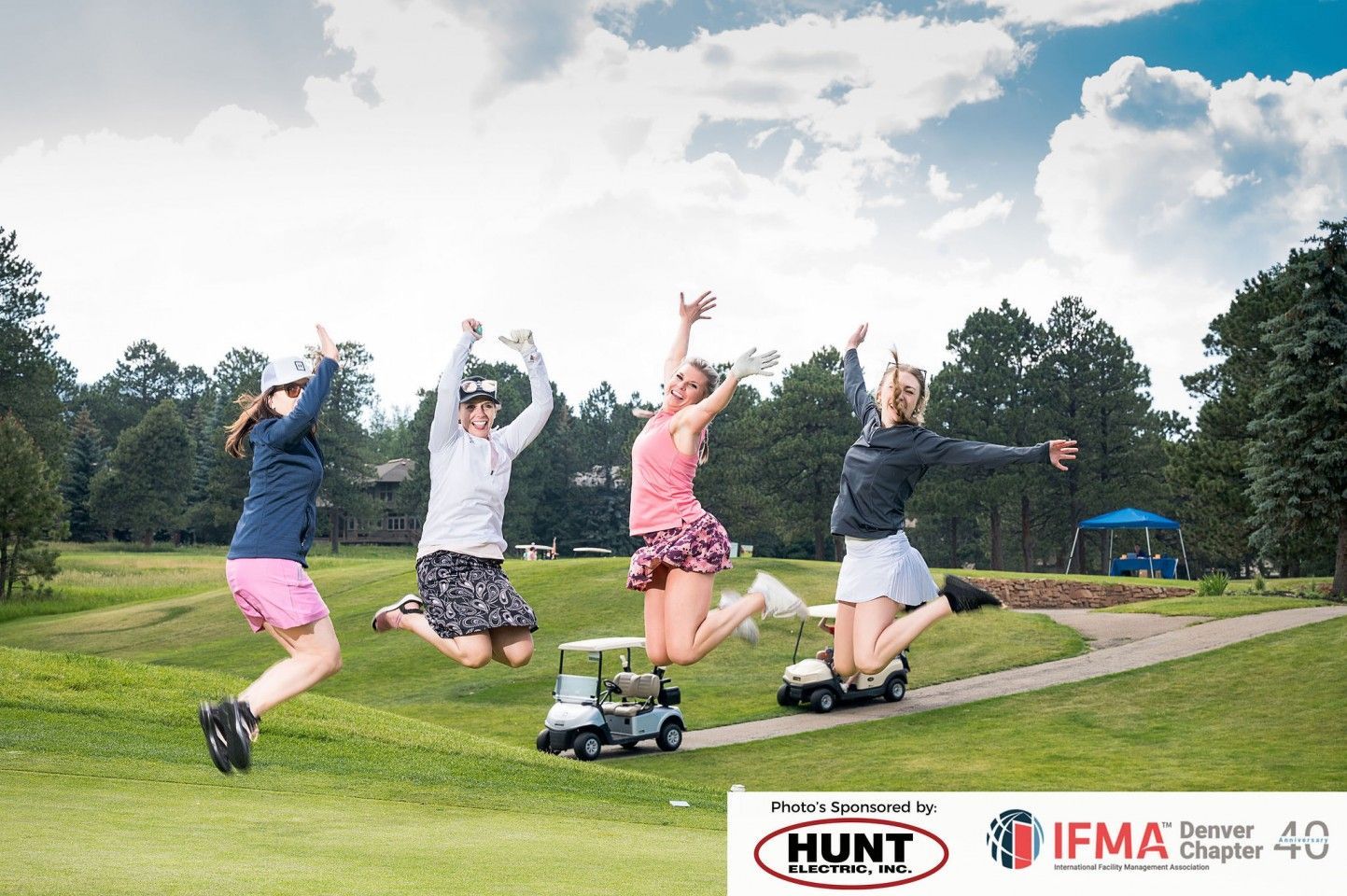 Four women golfers jump for joy on a golf course path next to golf carts.