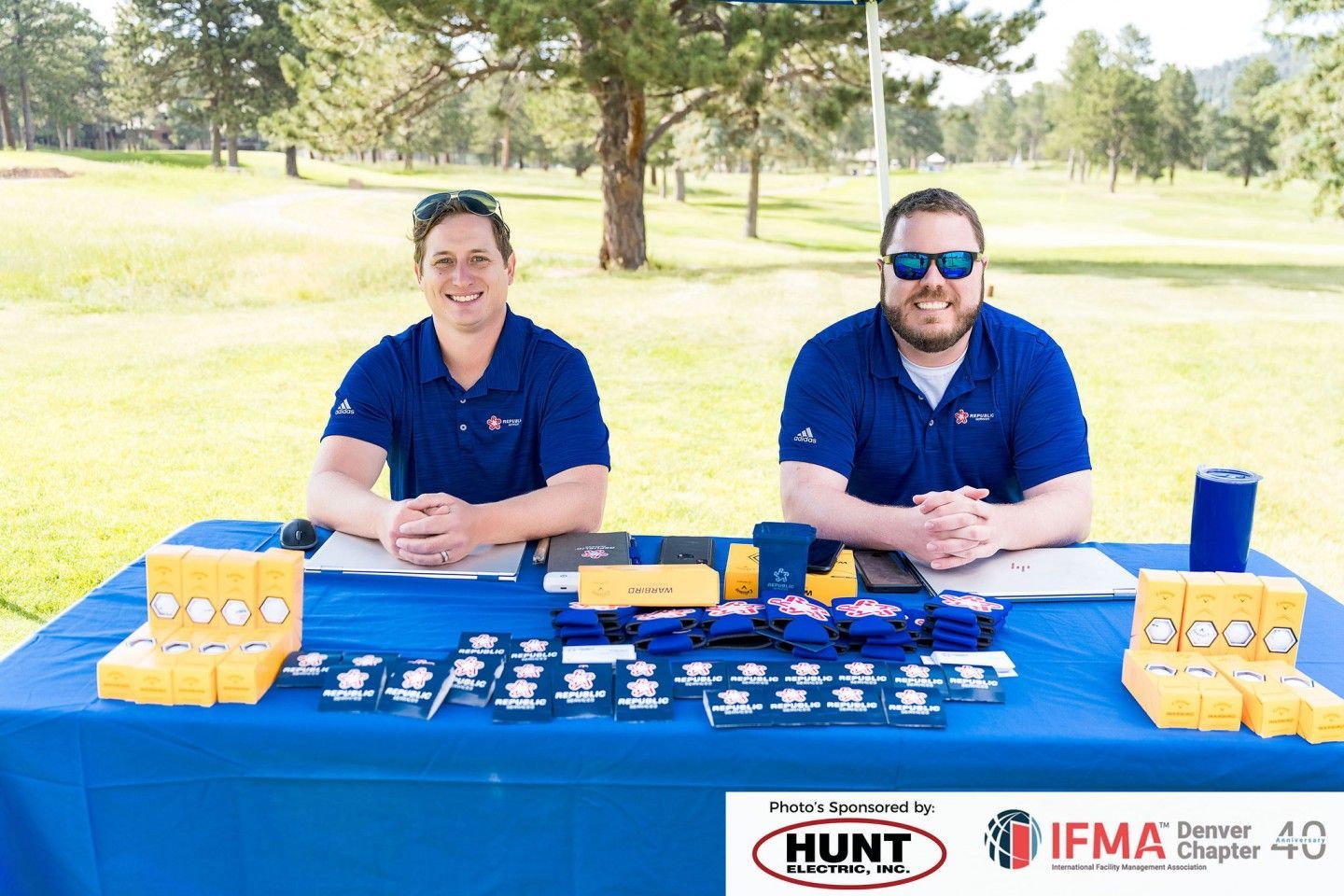 Two men at a blue table with promotional materials at an outdoor event.