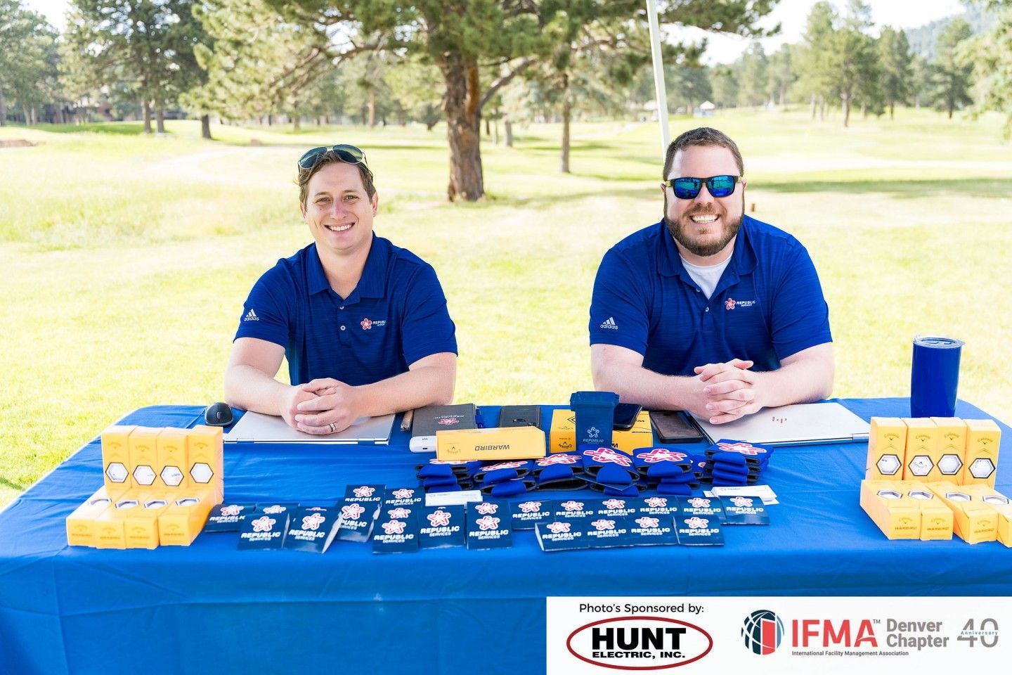 Two men at a table with promotional items at an outdoor event, wearing blue shirts. Blue table with branding.