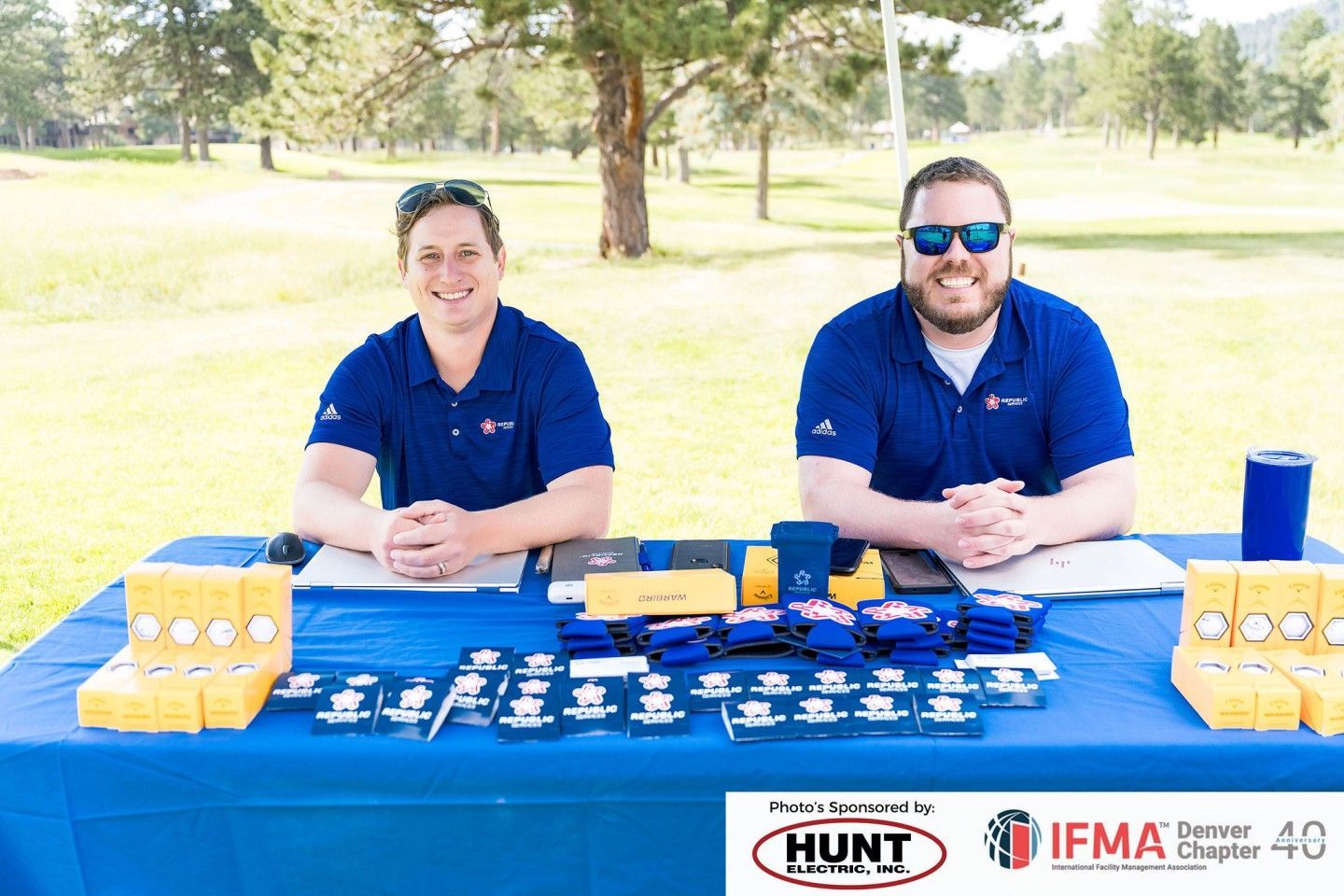 Two men at a table with promotional items, wearing blue shirts. Outdoor setting, blue table cover.
