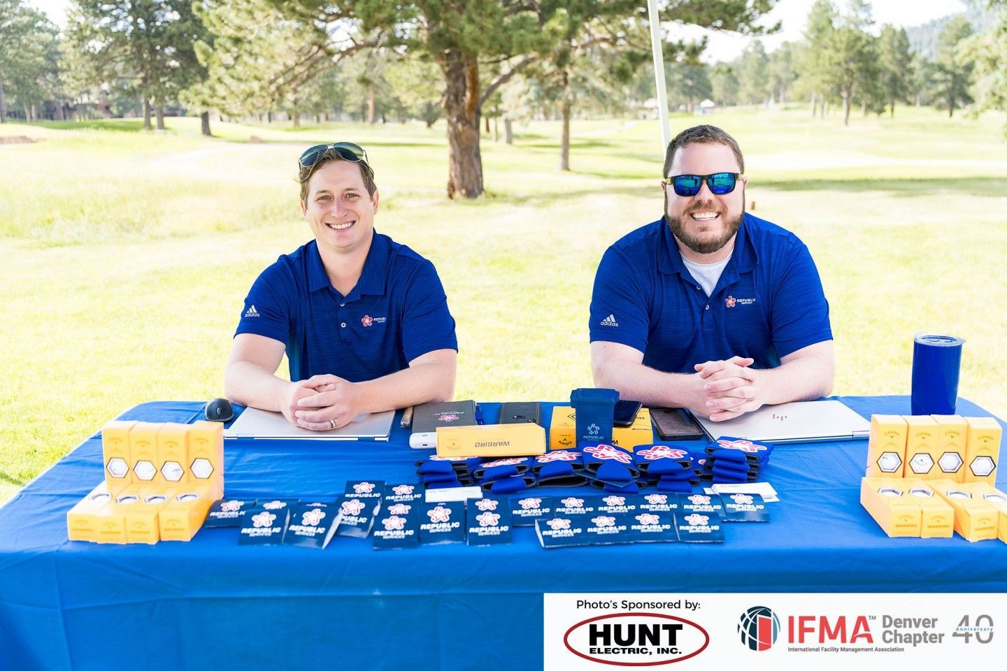 Two men at a blue table with promotional items outdoors. They wear blue shirts, smile. Sunlit green grass background.