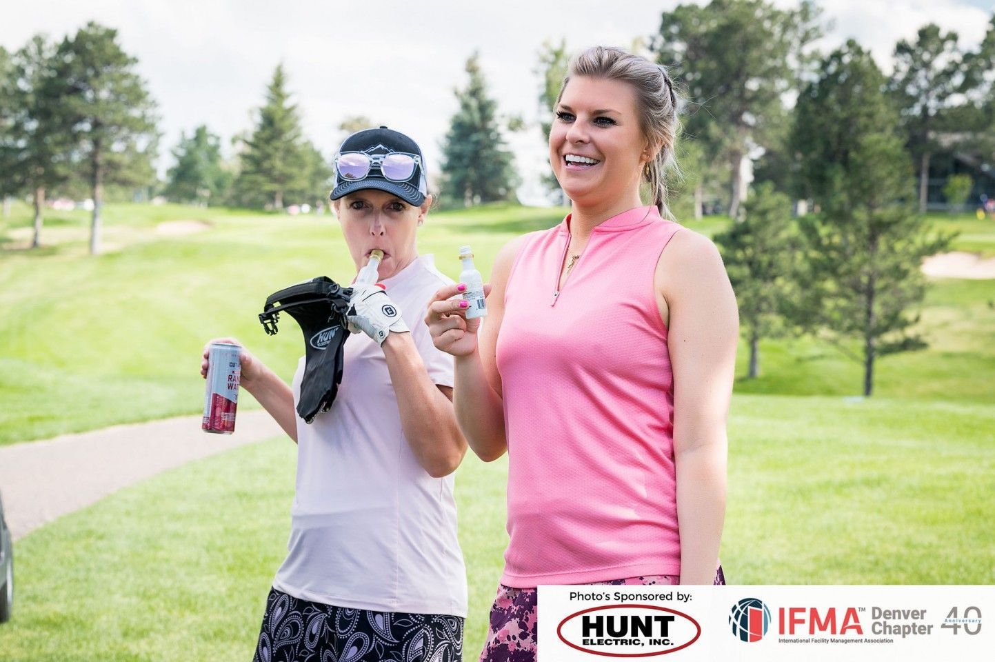 Two women on a golf course, one drinking a beverage, smiling, with a golf glove, trees in the background.