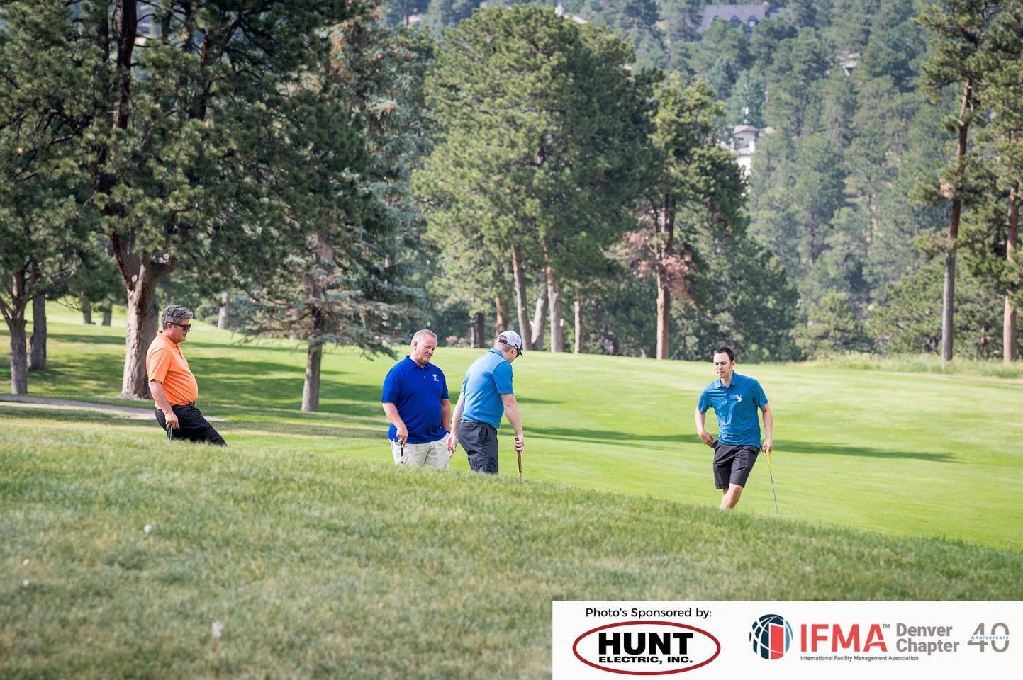 Men playing golf on a grassy course; trees and a hillside in the background.