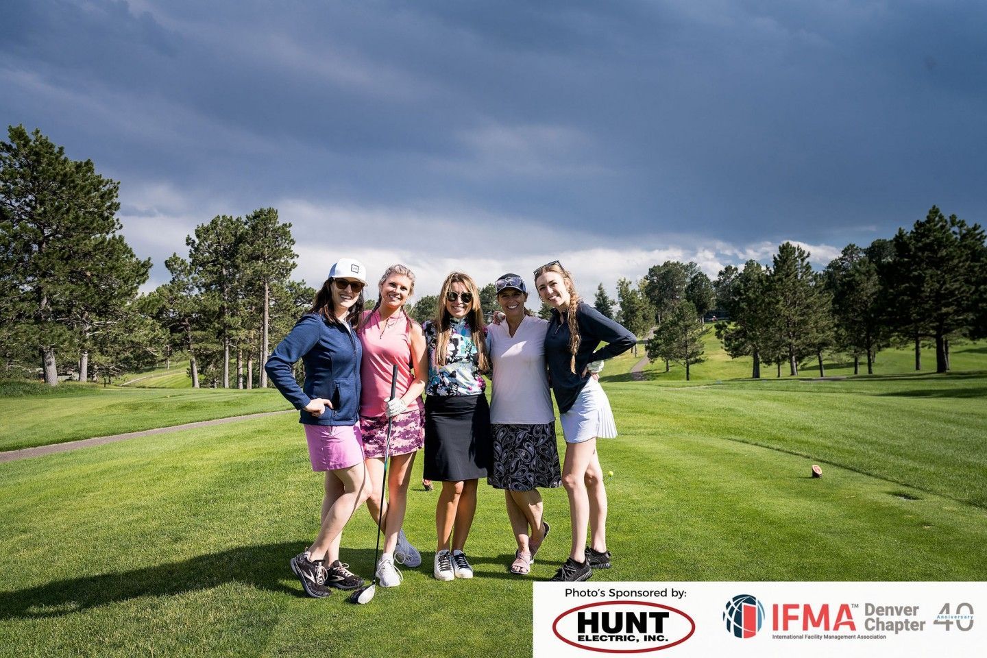 Six women smiling on a golf course. They are wearing golf attire, under a cloudy sky.