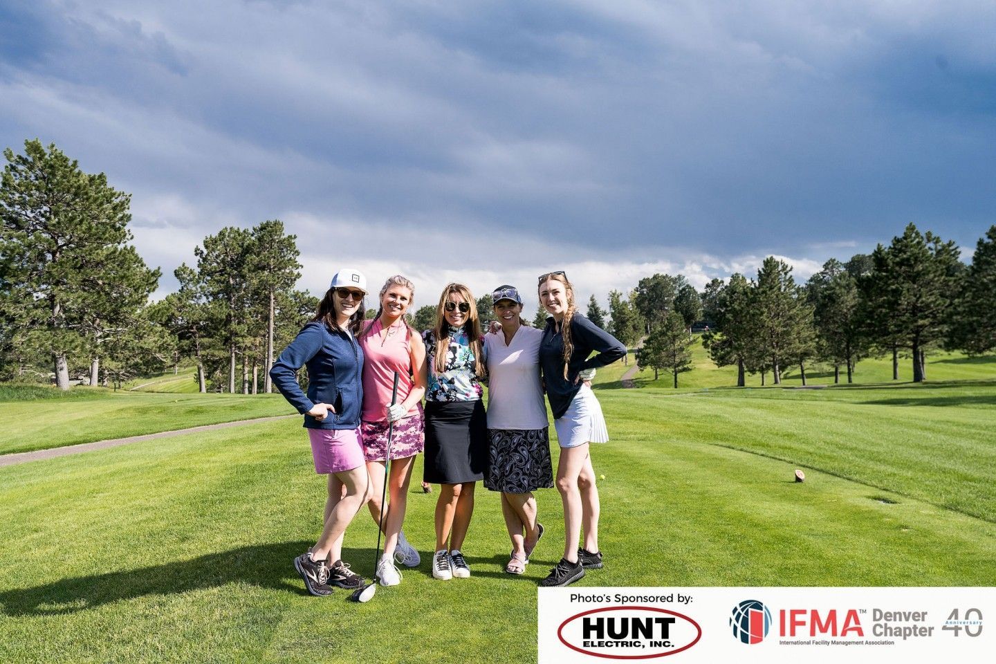 Five women stand on a golf course, posing for a photo. Overcast sky with trees in the background.