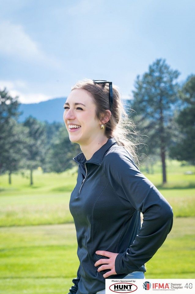Woman smiling on a golf course, wearing a gray shirt, with mountains and trees in the background.
