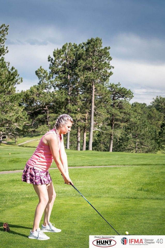 Woman golfing, poised to swing. Pink top, plaid skirt, on green fairway, trees in the background.