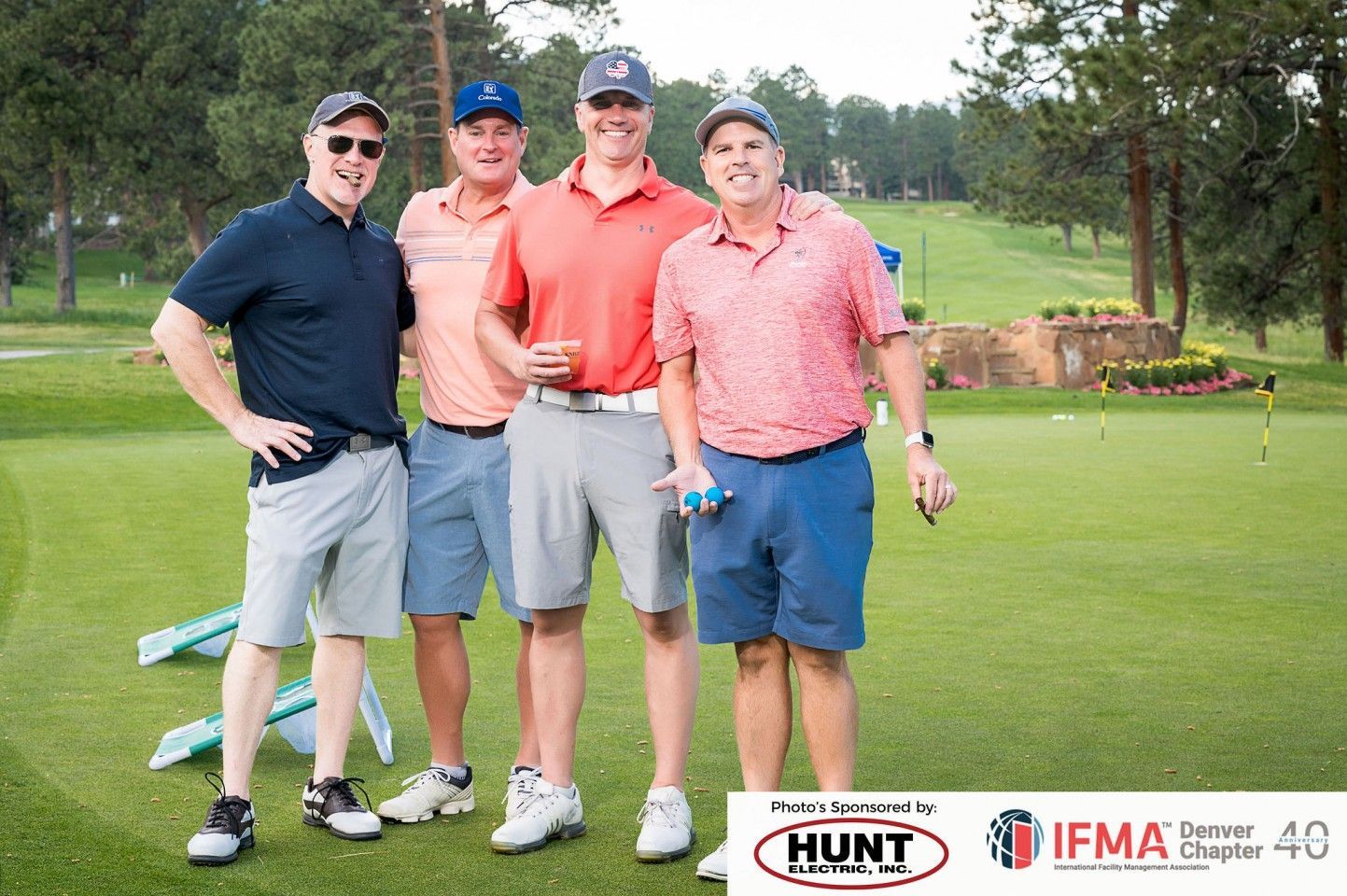 Four men in golf attire pose on a green, smiling. Golf course in background.
