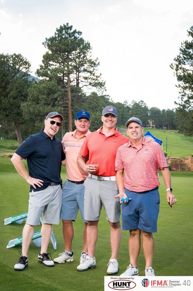 Four men in golf attire smile on a green course; trees in the background.