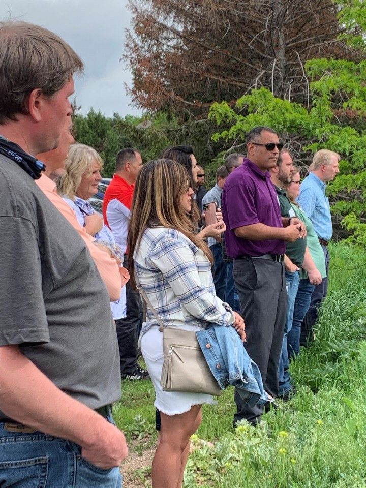 Group of people standing in a field, looking towards the right. Several wear casual clothing, some are smiling.