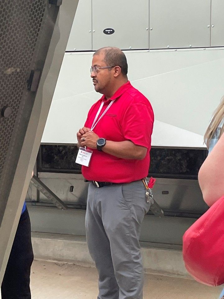 Man in red shirt and gray pants, looking left, near machinery; wearing name tag and smartwatch.