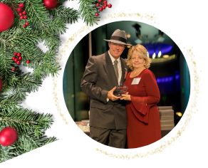 Man in suit and fedora with woman in red dress, holding award, holiday decor.