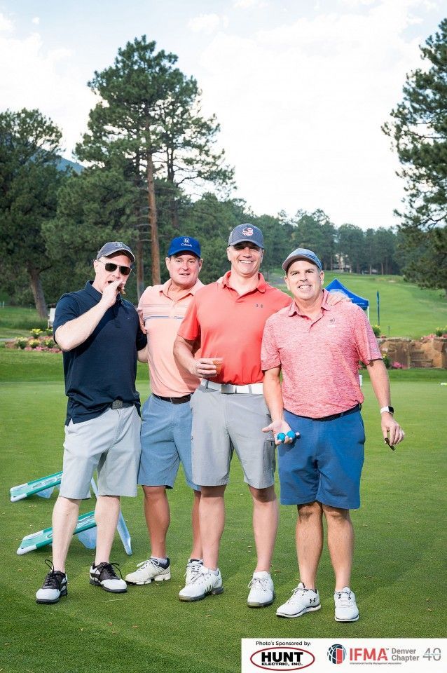 Four men smiling on a golf course. They are wearing hats, shorts and golf shirts. Green grass and trees in background.