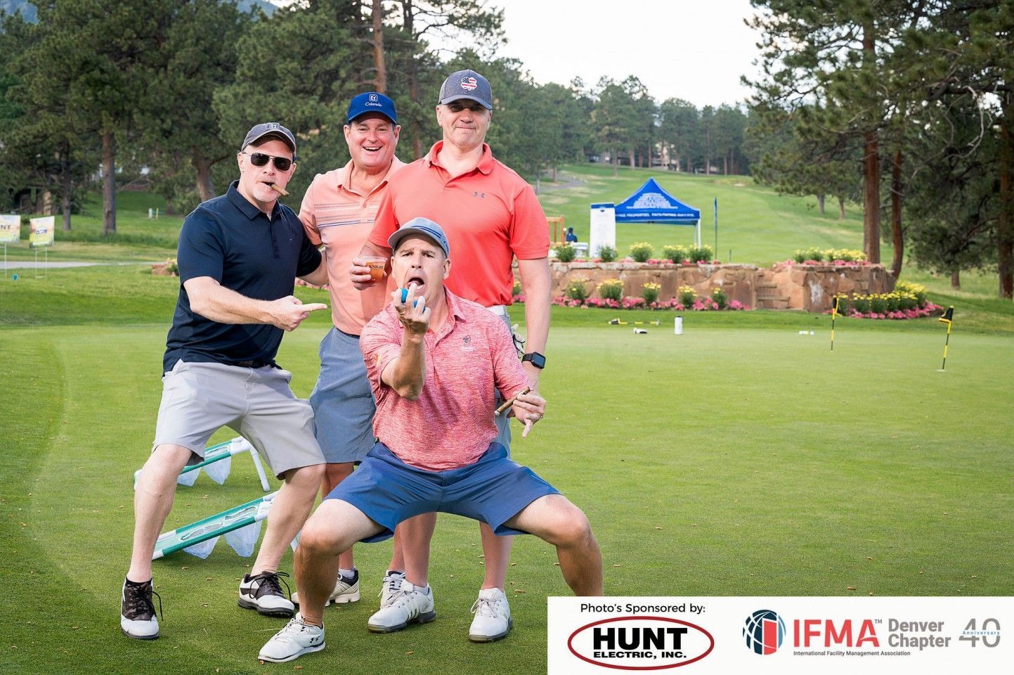 Four men pose on a golf course. One kneels with an open beverage, others point and smile.