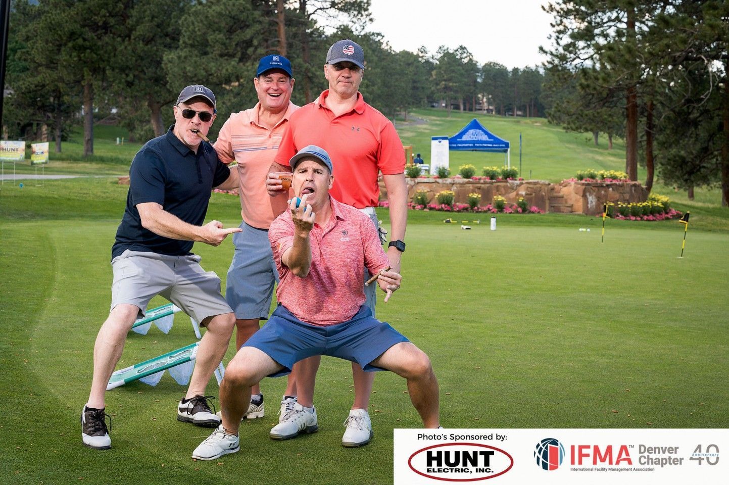 Four men on a golf course pose for a photo. One kneels, others stand, all with playful expressions.