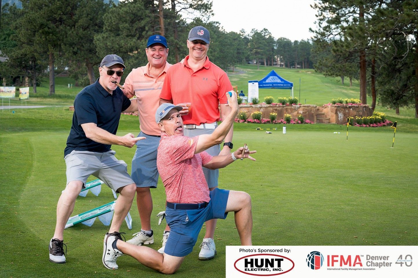 Four men pose on a golf course. One kneels and poses, others smile and gesture. Sunny day, green grass.