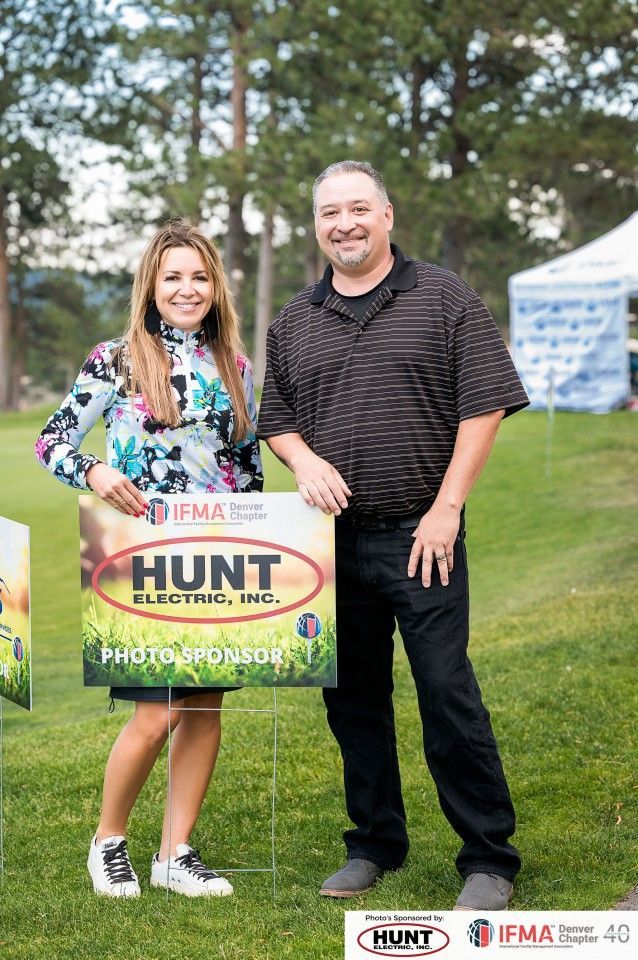 Two people stand on grass, holding a sign for Hunt Electric, photo sponsor.
