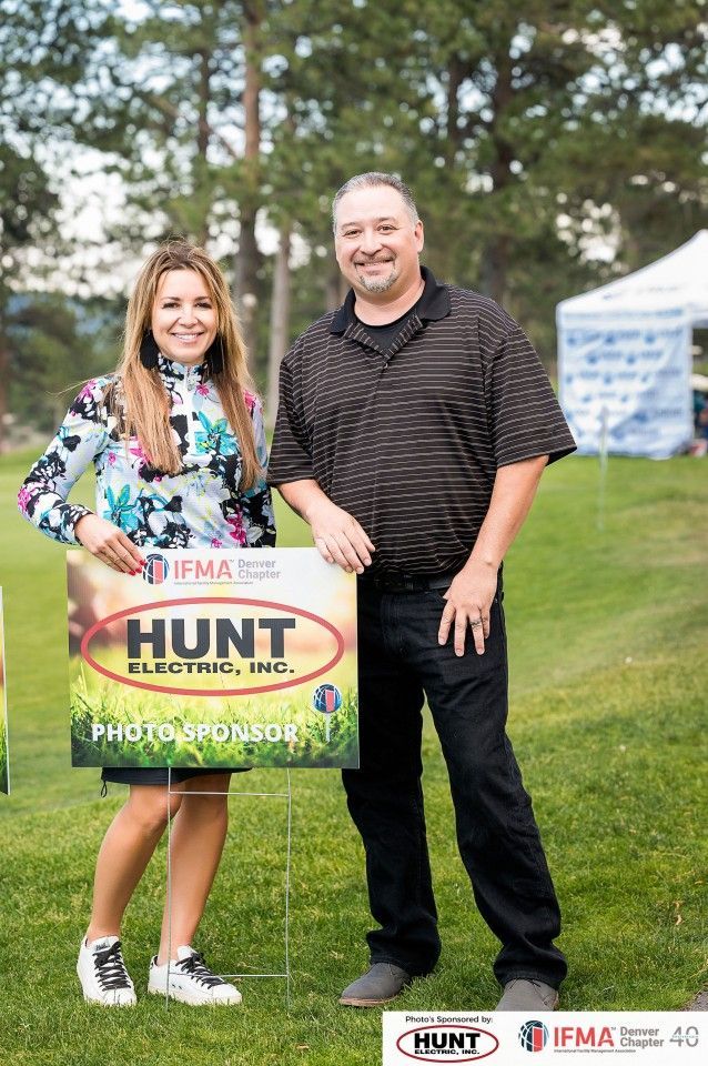 A woman and man stand on grass, holding a sign for Hunt Electric. Both smile.