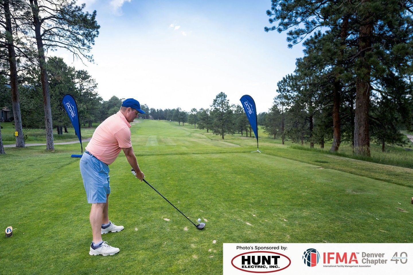 Golfer tees off on a green golf course, blue sky, trees in the background. Sponsored banners in view.