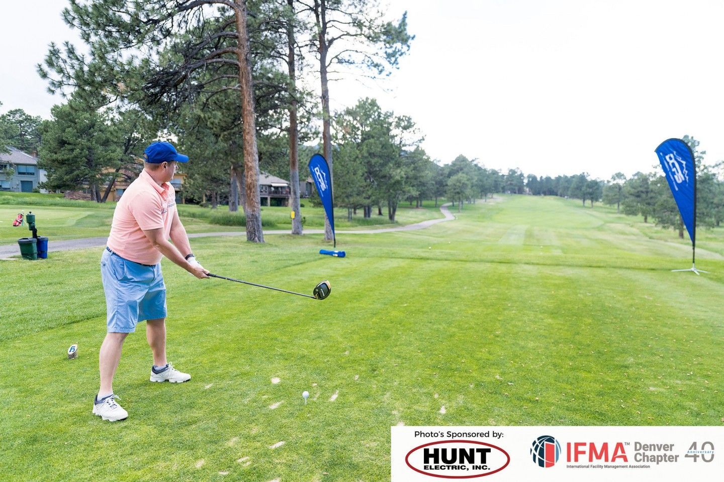 Man swings a golf club on a green course; blue banners and trees in the background.
