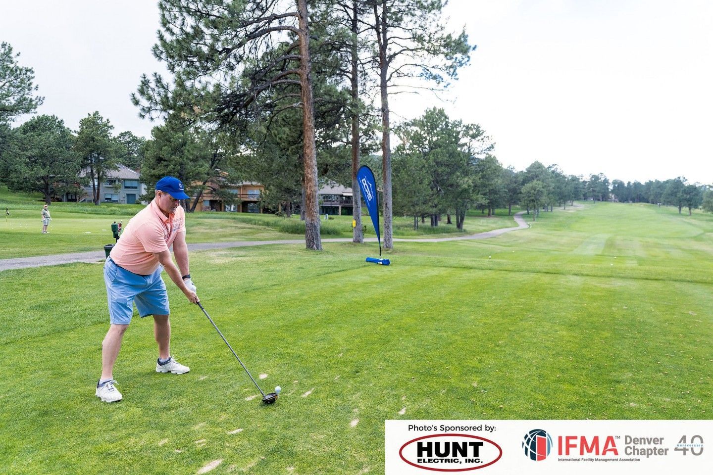 Man swings a golf club on a green course, teeing off near trees.