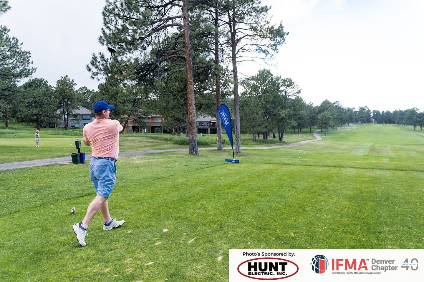 Man swings a golf club on a green course. Trees and buildings are in the background. Sponsors' logos are at the bottom.