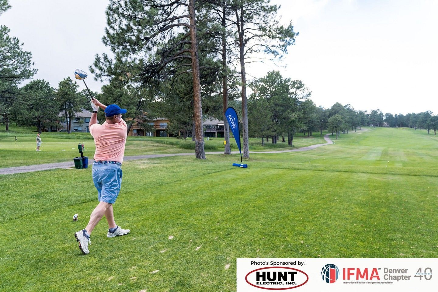 Golfer swinging club on a green course. Trees and buildings in background.