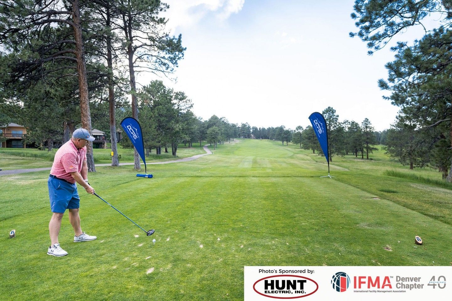 A golfer about to swing at a tee on a green golf course, blue sponsor flags present.