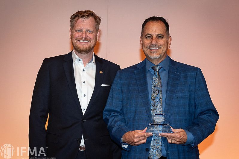 Two men smiling, one holding an award, orange and white background, formal attire.