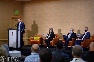 A speaker at a podium addresses a seated panel and audience in a conference room.