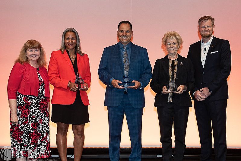 Five people stand on a stage holding awards. The background is a gradient of orange and white.