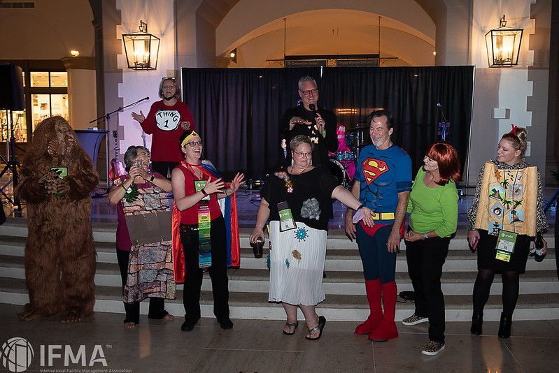 Group of people in costumes on a stage with a dark backdrop, some holding microphones, at a conference.