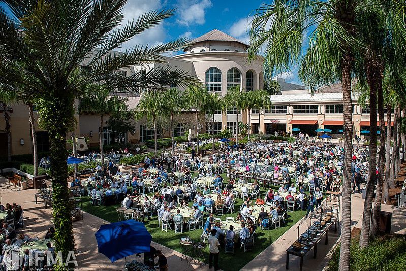 Large outdoor event; people seated at tables, palm trees, and a building with a rotunda.