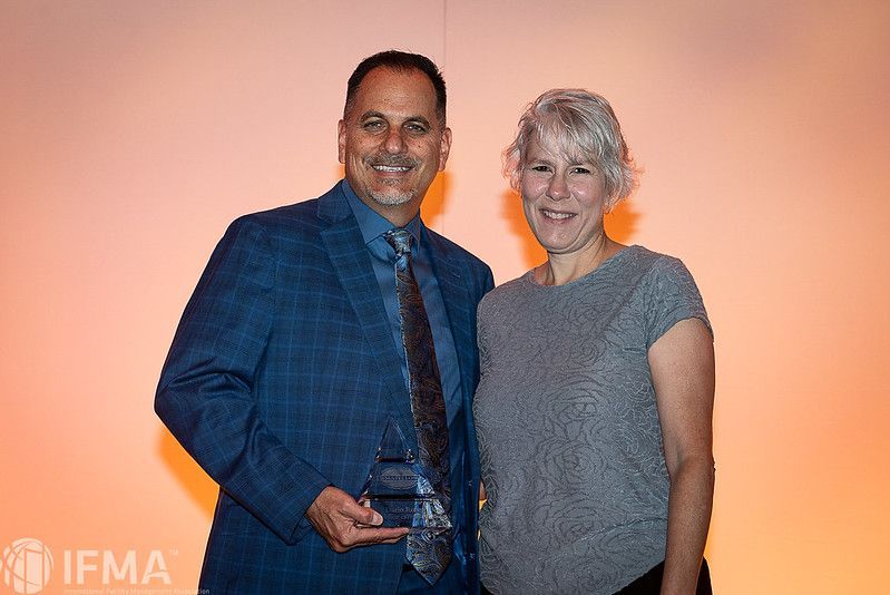 Man in blue suit holds award, woman in gray shirt stands beside him, orange background.