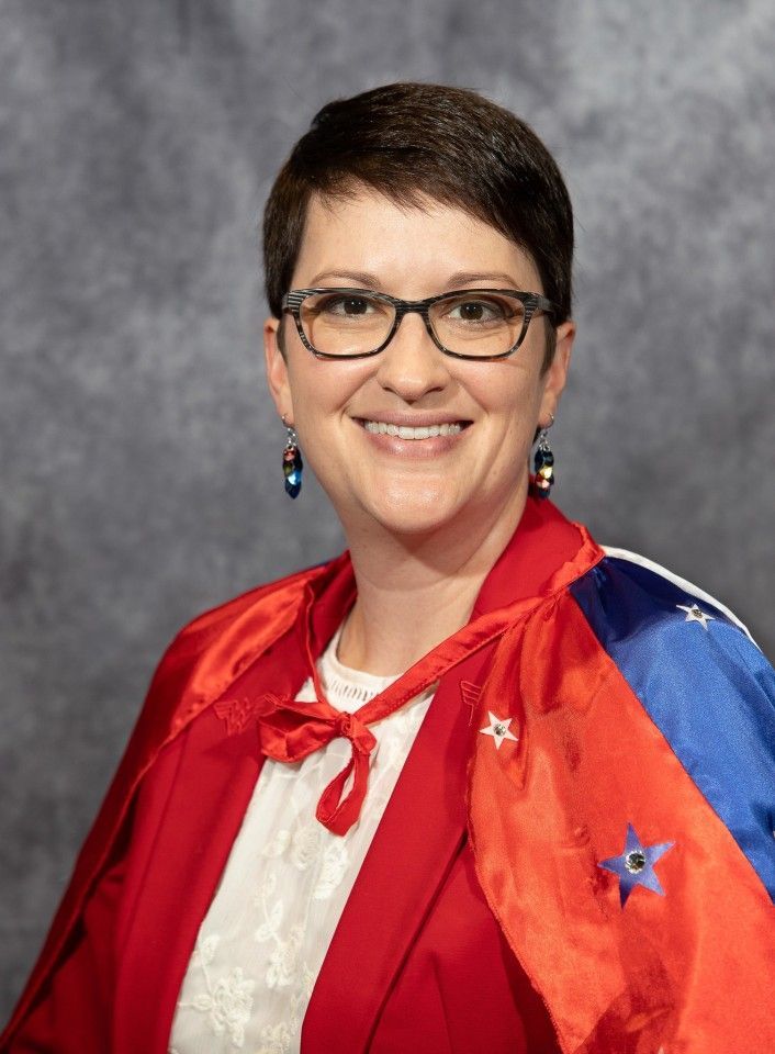 Woman with short hair, glasses, smiling, wearing a red and blue cape with stars, red jacket, white shirt.