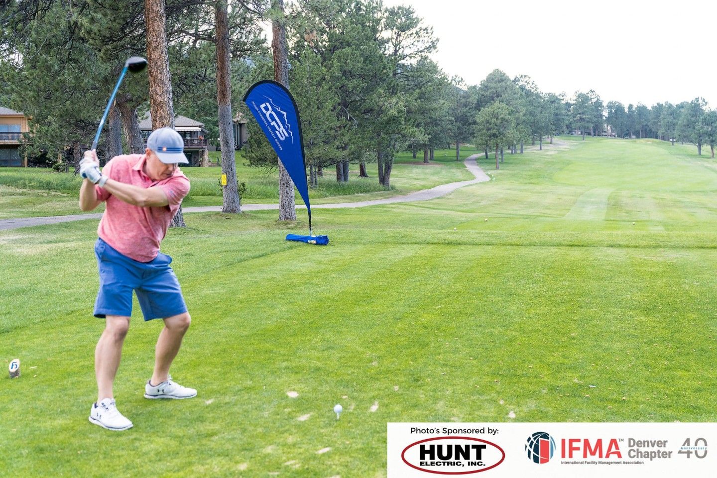 Golfer swinging a club on a green course; blue banner, trees, overcast day.
