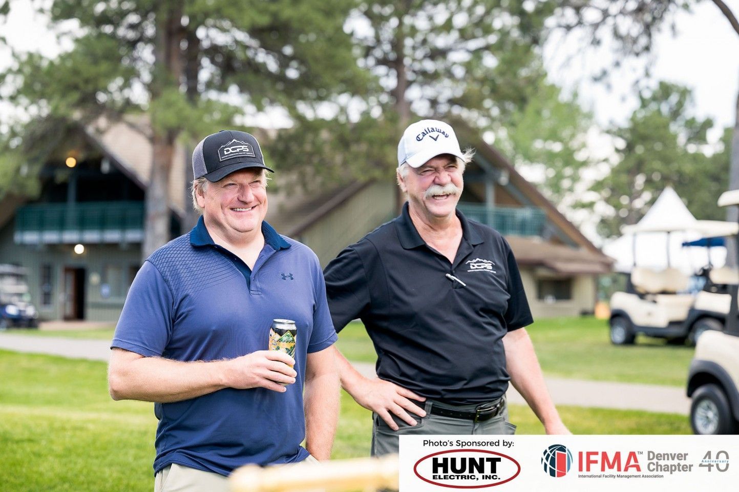 Two men smiling, one holding a beer, outside a building, near golf carts.