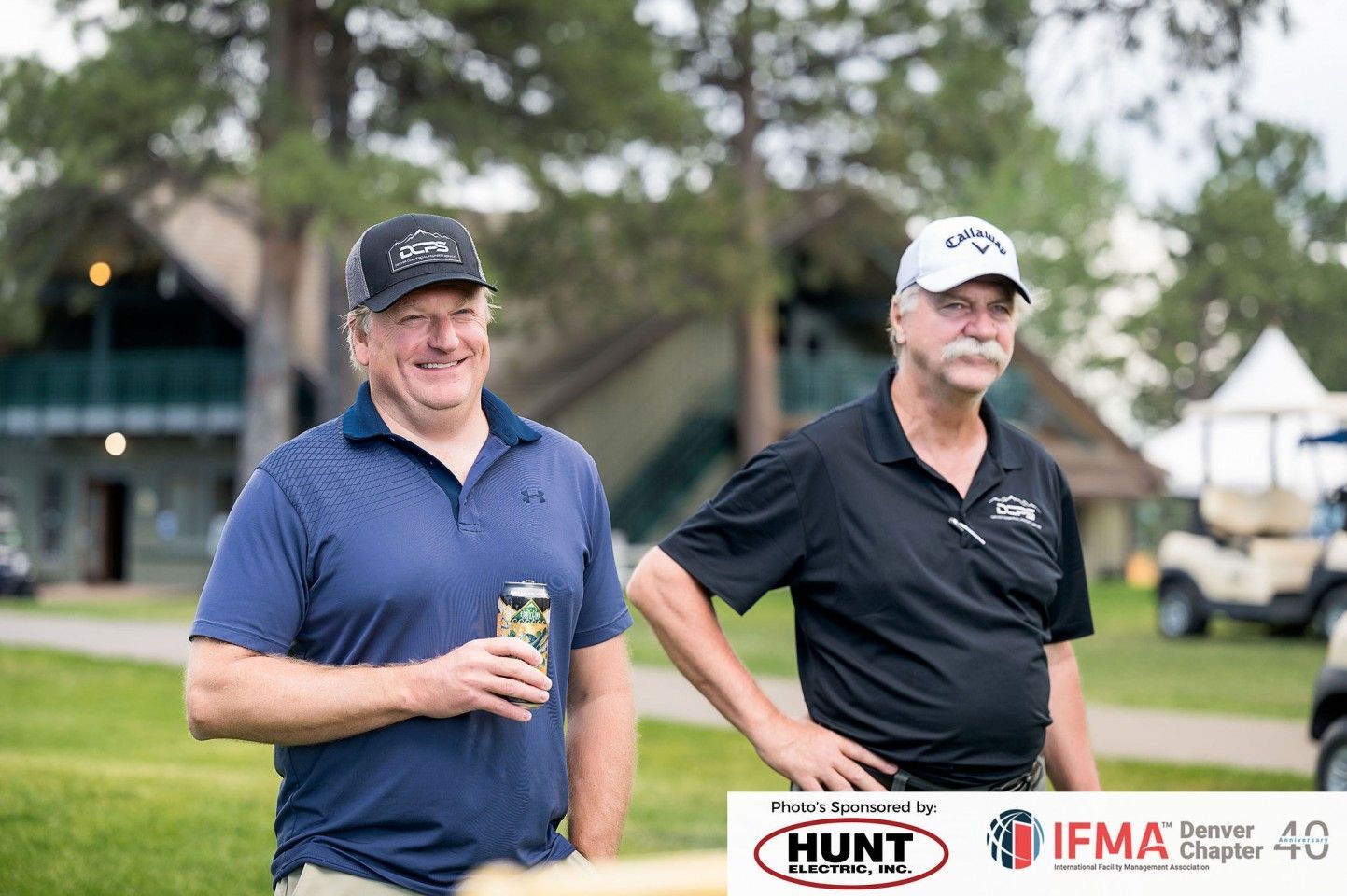 Two men at an outdoor event, one holding a drink, both wearing caps.