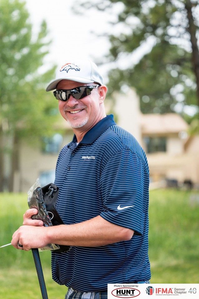 Man wearing sunglasses and a Broncos hat smiling, holding a golf club on a course.