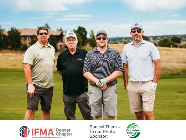 Four men pose on a golf course. They wear casual clothing, with a building and fields in the background.