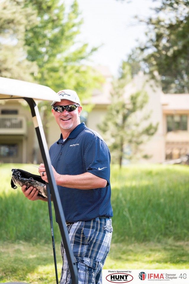 Man in golf attire, smiling, near a golf cart, holding a clipboard outside.