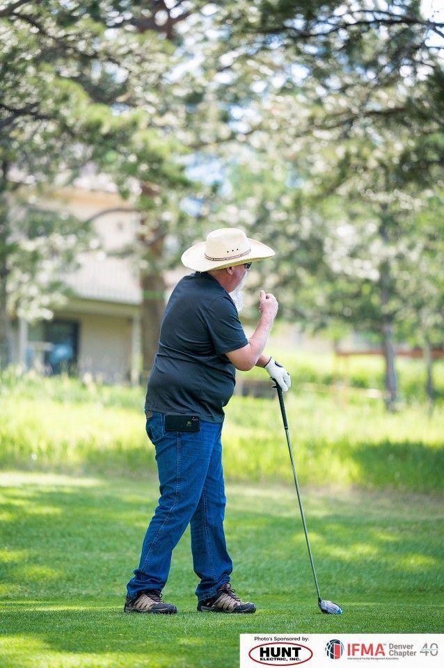 Man in straw hat and jeans on a golf course, holding a golf club, with trees in the background.