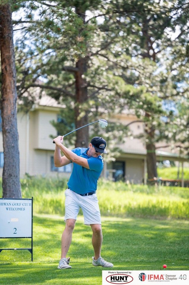 Man in blue shirt and white shorts swings a golf club on a green course, trees and building in background.