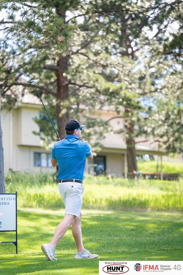 Man in blue shirt and white shorts swinging a golf club on a green course with a building in the background.