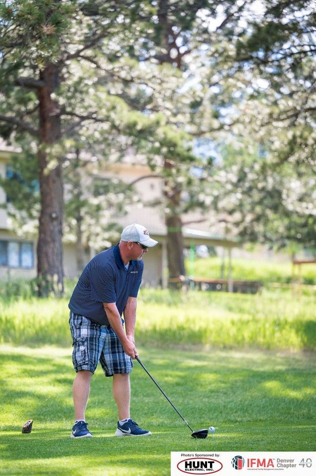 Man in golf attire swings a club on a green, trees and a building in the background.