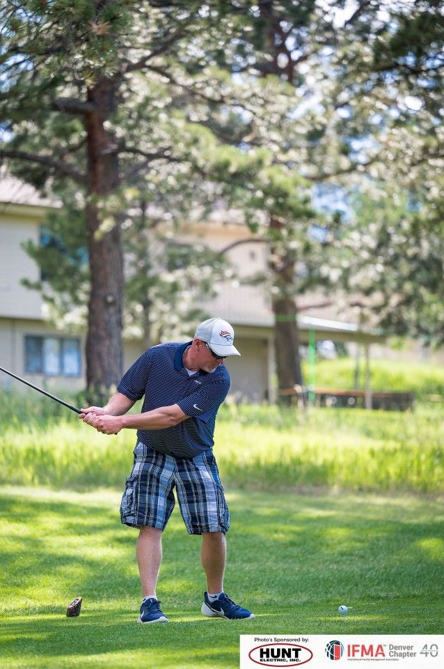 Man in plaid shorts swings golf club on a sunny course, tree and building in background.