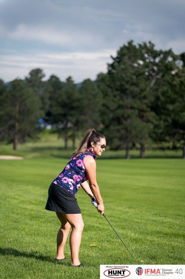 Woman swings a golf club on a green course, wearing a floral top and black skirt, with trees in the background.