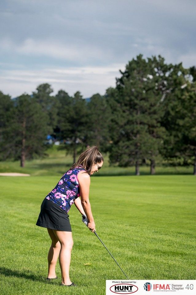 Woman in floral top and skirt golfing on a green course, trees and sky background.