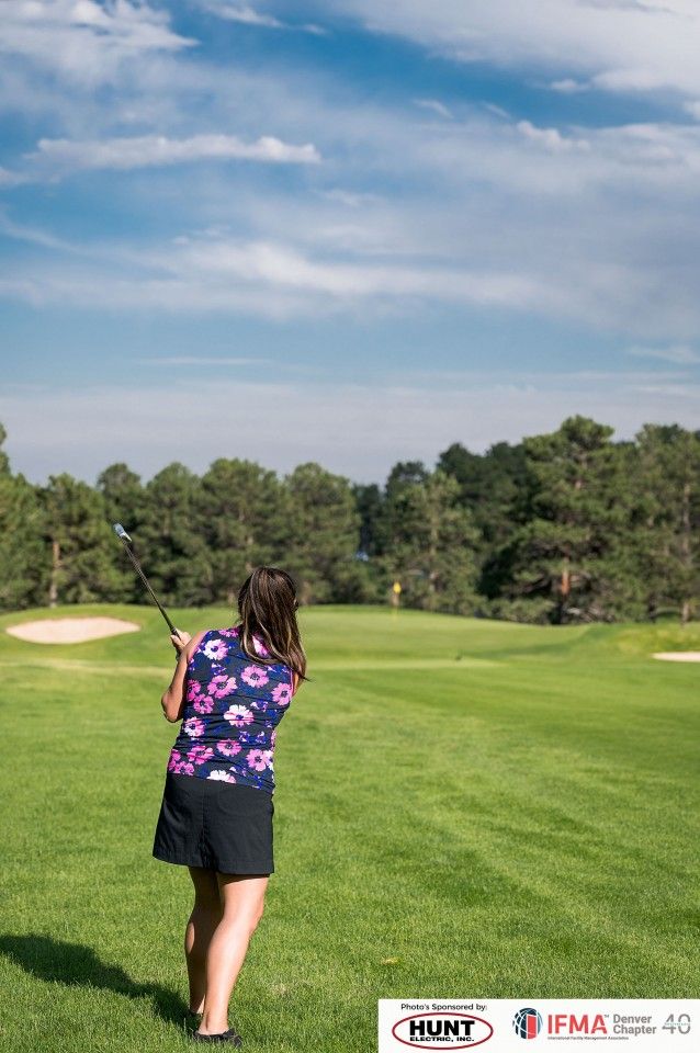 Woman swinging a golf club on a green course, with trees and a blue sky in the background.